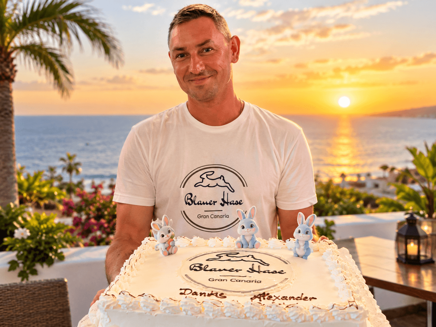 Dennis holds a cake decorated with the Blauer Hase logo in front of the sea at sunset